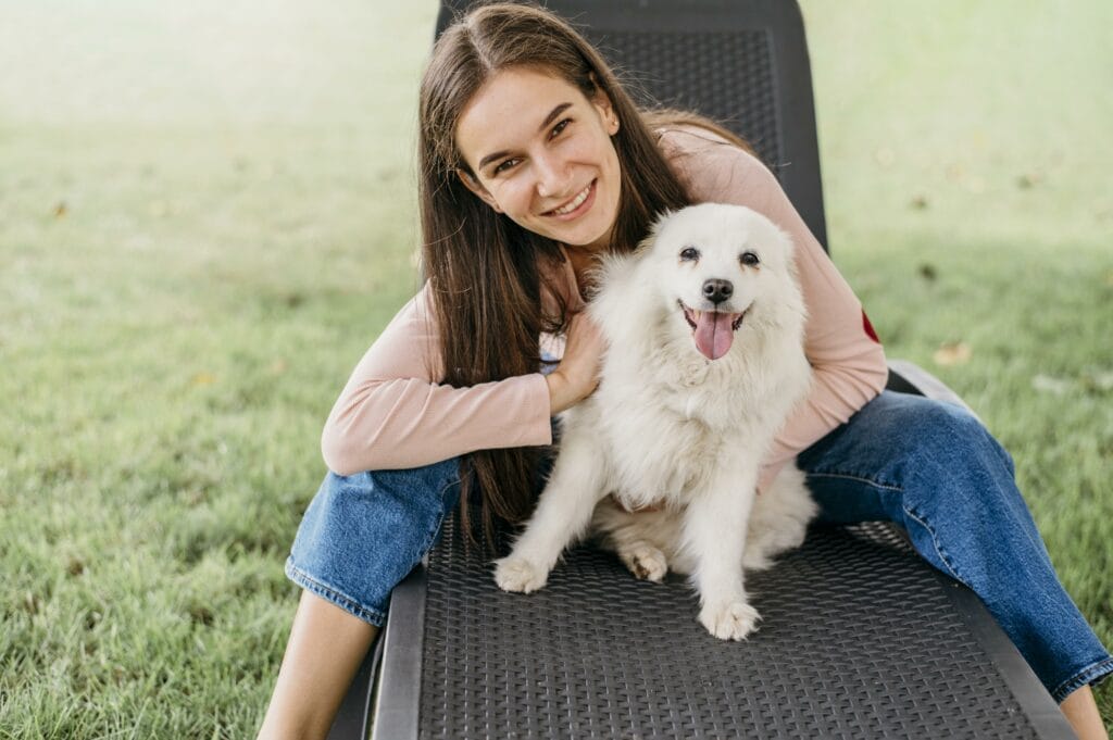 A young woman sits on a lounge chair outdoors, smiling and holding a small white dog with its tongue out, enjoying a sunny day after mobile dog grooming.