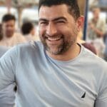 A man with dark hair and a beard, wearing a light blue shirt, smiles while sitting in a busy indoor setting—possibly chatting with friends about pet groomers NYC Queens NJ—with people in the background.