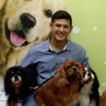 A man sits indoors with three Cavalier King Charles Spaniels; a large dog photo is displayed on the wall behind them, reminiscent of the style seen at top pet groomers NYC and Queens NJ residents trust.