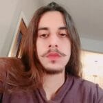 A person with long brown hair and a styled mustache and beard looks at the camera, wearing a purple shirt indoors—reminiscent of the artistic flair often seen among pet groomers NYC Queens NJ.