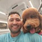 A man in a light blue shirt takes a selfie with his brown poodle inside a brightly lit room; the dog, just back from pet groomers NYC Queens NJ, is panting and looking towards the camera.