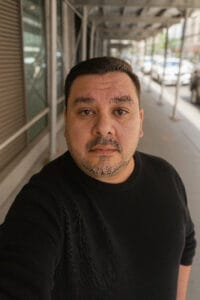 A man with short dark hair and a trimmed beard wears a black sweater and stands on a city sidewalk under scaffolding in Queens, home to some of the top pet groomers NYC and NJ have to offer, looking directly at the camera.